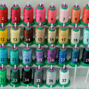 May include: A set of 37 spools of thread, arranged in rows on a white shelf. The spools are numbered 1 to 37. The thread colours include red, yellow, blue, green, purple, black, white, and beige.