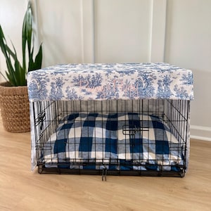 May include: A black wire dog crate with a blue and white toile fabric cover and a blue and white plaid dog bed inside. The fabric cover features a scenic design with trees and figures. The dog bed is a square shape.