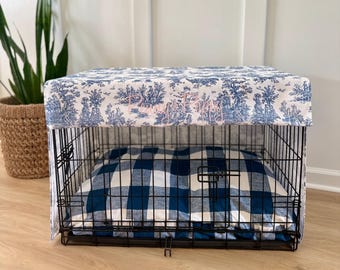 May include: A black wire dog crate with a blue and white toile fabric cover and a blue and white plaid dog bed inside. The fabric cover has a scenic design with trees and figures. The dog bed is a square shape.