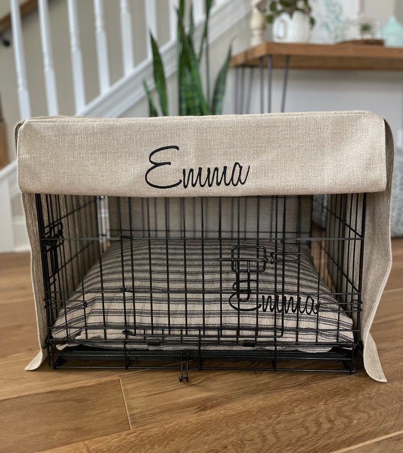 May include: A beige linen dog crate cover with the name "Emma" embroidered on the front. The cover is on a black metal dog crate with a striped bed inside.