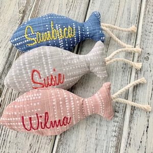 May include: Three fabric fish toys with embroidered names. The fish are gray, blue, and pink with white polka dots. The names are "Sambuca", "Susie", and "Wilma".