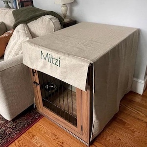 May include: A beige linen dog crate cover with the name "Mitzi" embroidered on the front. The cover is draped over a wooden crate with a black metal wire door.