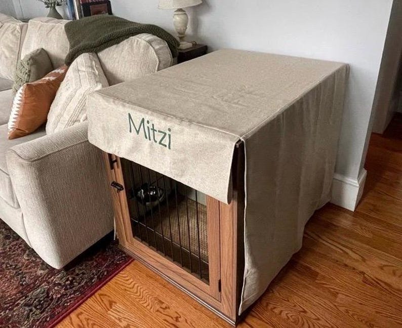 May include: A beige linen dog crate cover with the name "Mitzi" embroidered on the front. The cover is draped over a wooden crate with a black metal wire door.