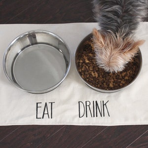 May include: A white cotton dog mat with the words "EAT" and "DRINK" embroidered on it. Two stainless steel bowls are on the mat, one with dog food and a small dog is eating from it.