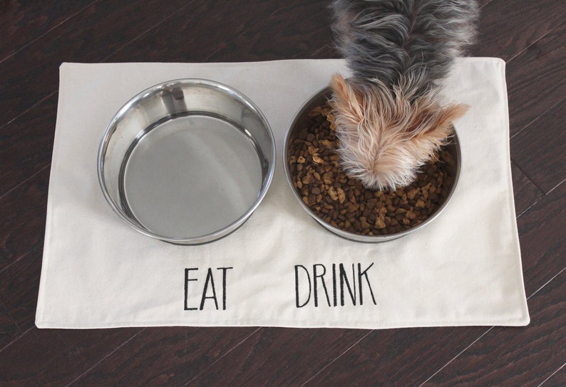 May include: A white cotton dog mat with the words "EAT" and "DRINK" embroidered on it. Two stainless steel bowls are on the mat, one with dog food and a small dog is eating from it.