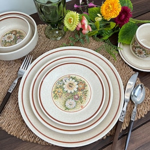 May include: A vintage cream-colored dinnerware set with a floral design in the center of each piece. The set includes plates, bowls, and a cup and saucer. Brown trim borders the plates. Silverware with dark brown handles is also present.