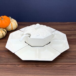 May include: A white ceramic serving set with a central lidded bowl and a matching spoon, surrounded by six octagonal serving dishes. The bowl and lid feature a leaf design. The set is on a wooden surface.