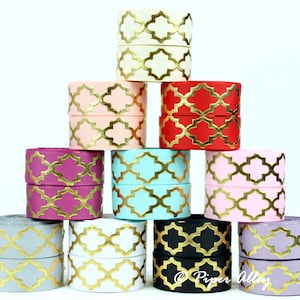 May include: A collection of stacked ribbon spools in various colors, including cream, pink, red, purple, turquoise, gray, black, and lavender. Each spool features a gold quatrefoil pattern. The spools are arranged in a pyramid shape against a white background.