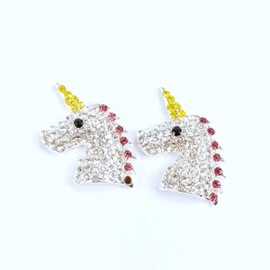 May include: Two silver-toned unicorn head-shaped earrings. Each earring is encrusted with clear rhinestones, with a yellow horn, black eye, and red mane. The earrings are set against a white background.