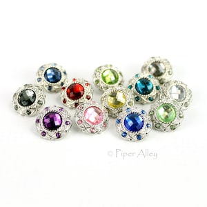 May include: A collection of decorative buttons with jewel-toned center stones in various colors, including blue, red, and green. Each button has a silver-colored, ornate metal frame. The buttons are arranged on a white background.