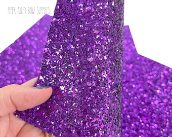 May include: Close-up of a sheet of vibrant purple glitter fabric. The fabric is densely covered in sparkling glitter particles, reflecting light. The text "Piper Alley Bow Supplies" is visible in the upper left corner. The fabric is being held by a hand.
