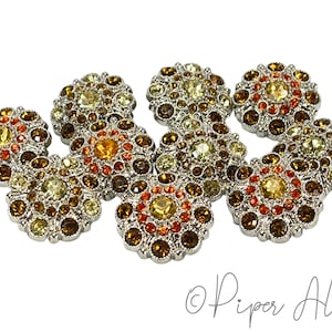 May include: A collection of eight decorative buttons with a floral design. Each button features a silver-colored metal base and is adorned with a variety of amber, orange, and yellow rhinestones. The buttons are arranged on a white background.