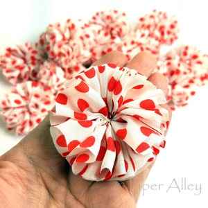 May include: A close-up of a white fabric flower with red polka dots. The flower is made of multiple layers of fabric and is gathered in the center. The flower is being held in a hand.