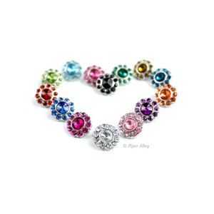 May include: A collection of colorful, flower-shaped snap jewelry components arranged in a heart shape. The assortment includes various colors like blue, pink, green, and orange, each with a central gem surrounded by smaller stones. The background is white.