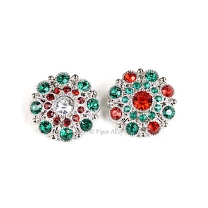 May include: Two silver-toned, round snap jewelry pieces with a floral design. Each piece features a central gemstone surrounded by smaller red and green gems. The gems are set in a silver metal base.
