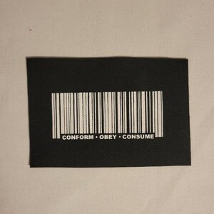 Puede incluir: Parche negro con un código de barras blanco y el texto "CONFORM. OBEY. CONSUME."