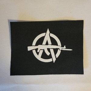 Może przedstawiać: Czarna prostokątna naszywka z białym symbolem anarchii. Symbol zawiera okrąg, literę A i karabin. Naszywka jest prawdopodobnie wykonana z tkaniny i jest przeznaczona do przyszywania lub naprasowywania na odzież lub akcesoria.