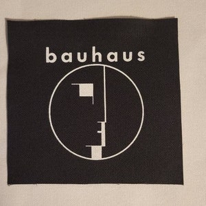Peut inclure: Patch thermocollant noir et blanc avec le nom du groupe "bauhaus" et un graphique de visage stylisé.
