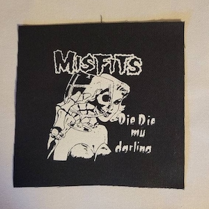 May include: A black and white patch with the band name "Misfits" in a stylised font. The patch features a black and white illustration of a woman with a skull for a face holding a glass of wine. The text "Die Die mu darling" is below the illustration.