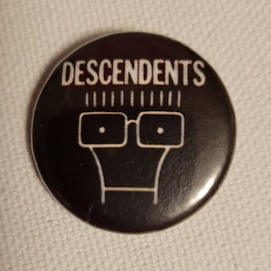 Könnte beinhalten: Schwarz-weißer Knopf mit dem Bandnamen "DESCENDENTS" in weißer Schrift und einer stilisierten Illustration einer Person mit Brille.