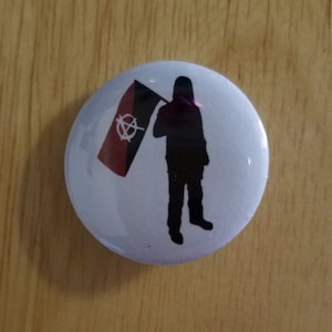 Puede incluir: Un botón blanco con una silueta negra de una persona sosteniendo una bandera roja y negra con un símbolo de anarquía blanco.