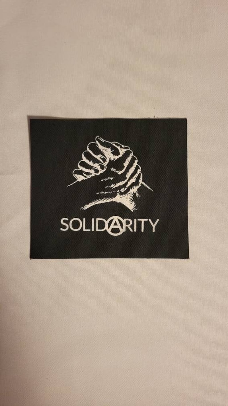 Puede incluir: Parche negro con una imagen blanca de dos manos entrelazadas y la palabra "SOLIDARITY" con un s&iacute;mbolo de anarqu&iacute;a en lugar de la "A".