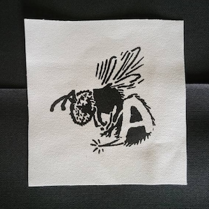 May include: Black and white embroidered patch of a bee with the letter "A" on its back. The bee is flying with its wings spread out. There is a small spark of light under the bee.