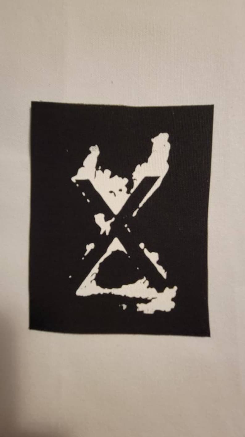 Op de afbeelding: Een zwarte stoffen patch met een witte gestileerde letter X.