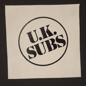 Puede incluir: Un parche de tela blanco con un círculo negro que contiene el texto "U.K. SUBS" en una fuente negrita y en bloque.