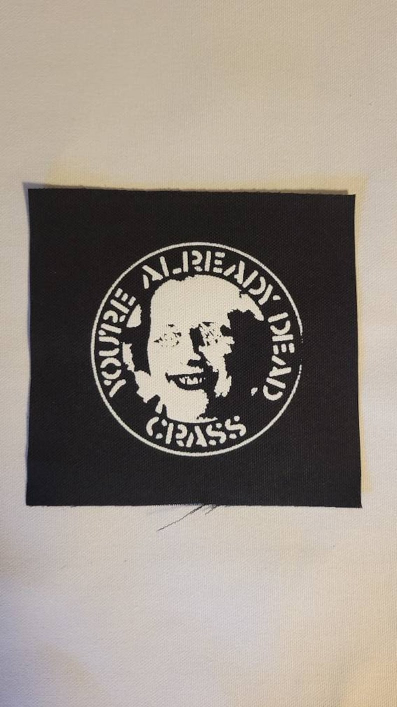 May include: A black and white patch with a circle design. The circle features a white image of a person's face with the text "YOU'RE ALREADY DEAD" and "CRASS" around the image.