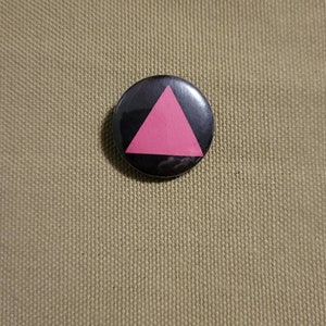 Pode incluir: Um botão preto e rosa com um triângulo rosa no centro.