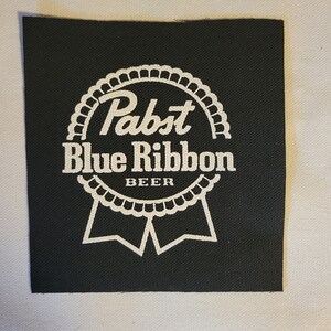 Può includere: Toppa in tessuto nero con un logo bianco Pabst Blue Ribbon Beer. Il logo presenta un nastro bianco con le parole "Pabst Blue Ribbon Beer" in un font stilizzato.