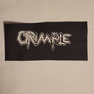 Könnte beinhalten: Ein schwarzer Stoff-Patch mit dem Wort "CRUMPLE" in weißer, stilisierter Schrift.