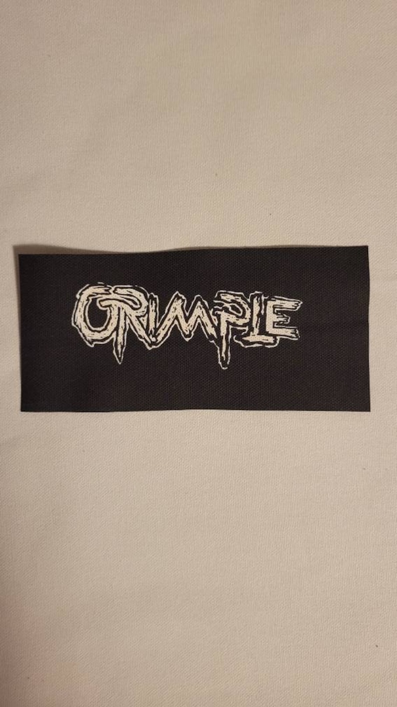 Op de afbeelding: Een zwarte stoffen patch met het woord "CRUMPLE" in witte, gestileerde letters.