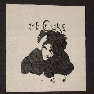 Op de afbeelding: Een witte stoffen patch met zwarte inkt die een silhouet van een persoon afbeeldt met de tekst "THE CURE" boven de persoon.