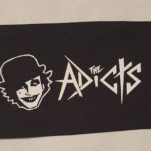 May include: Black and white patch with the text "The Adicts" in a stylized font. A cartoon face with a hat is on the left side of the patch.