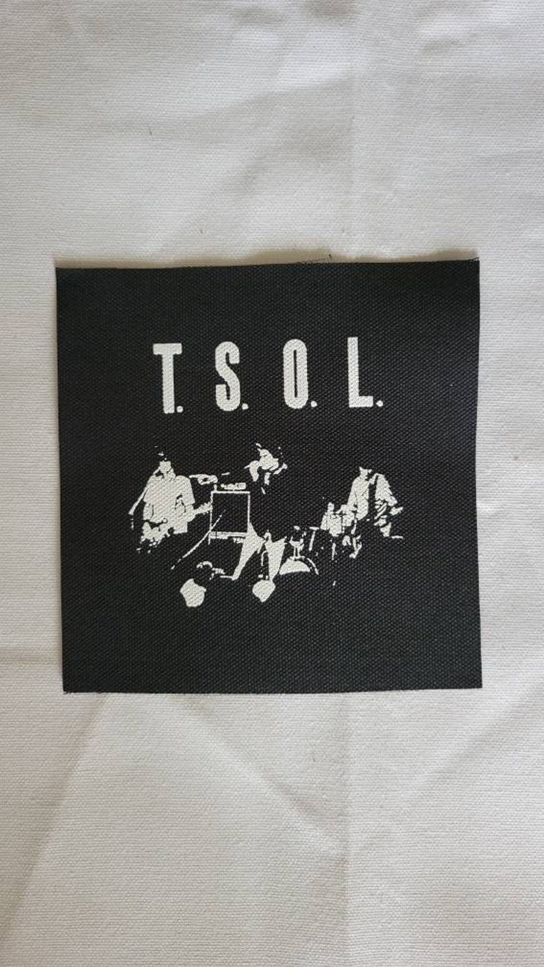 Puede incluir: Parche blanco y negro con el texto "T.S.O.L." en letras blancas. El parche presenta una silueta de una banda tocando m&uacute;sica.