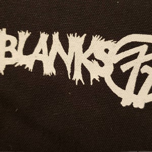 May include: Black fabric with a white graphic. The graphic features the word "BLANKS" in a jagged font, and the number "77" inside a circle. The design has a punk rock or alternative aesthetic.