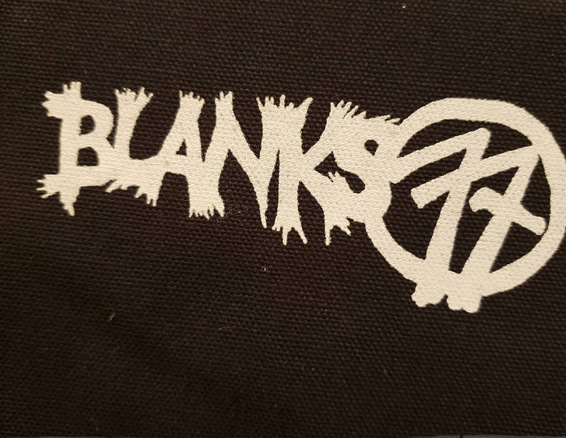 May include: Black fabric with a white graphic. The graphic features the word "BLANKS" in a jagged font, and the number "77" inside a circle. The design has a punk rock or alternative aesthetic.