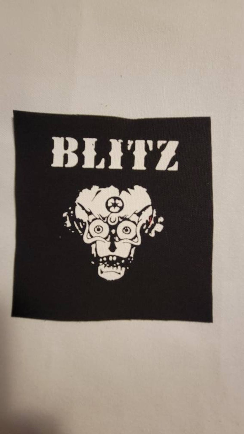 May include: Black and white patch with the word "BLITZ" in white letters and a white illustration of a skull with a third eye in the center of the forehead.