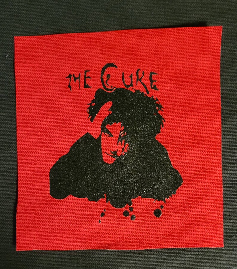 Puede incluir: Parche termoadhesivo en blanco y negro con una silueta de una persona con el texto "THE CURE" sobre la imagen. El parche est&aacute; sobre un fondo rojo.