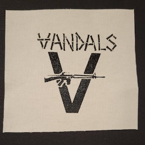 May include: Square, off-white patch with the word "VANDALS" in a stencil font above a large black "V". A rifle is horizontally centered within the "V". The patch is on a black background.