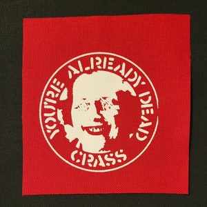 May include: A red fabric patch with a white circle containing a stylized image of a person's face and the text "YOU'RE ALREADY DEAD, CRASS".