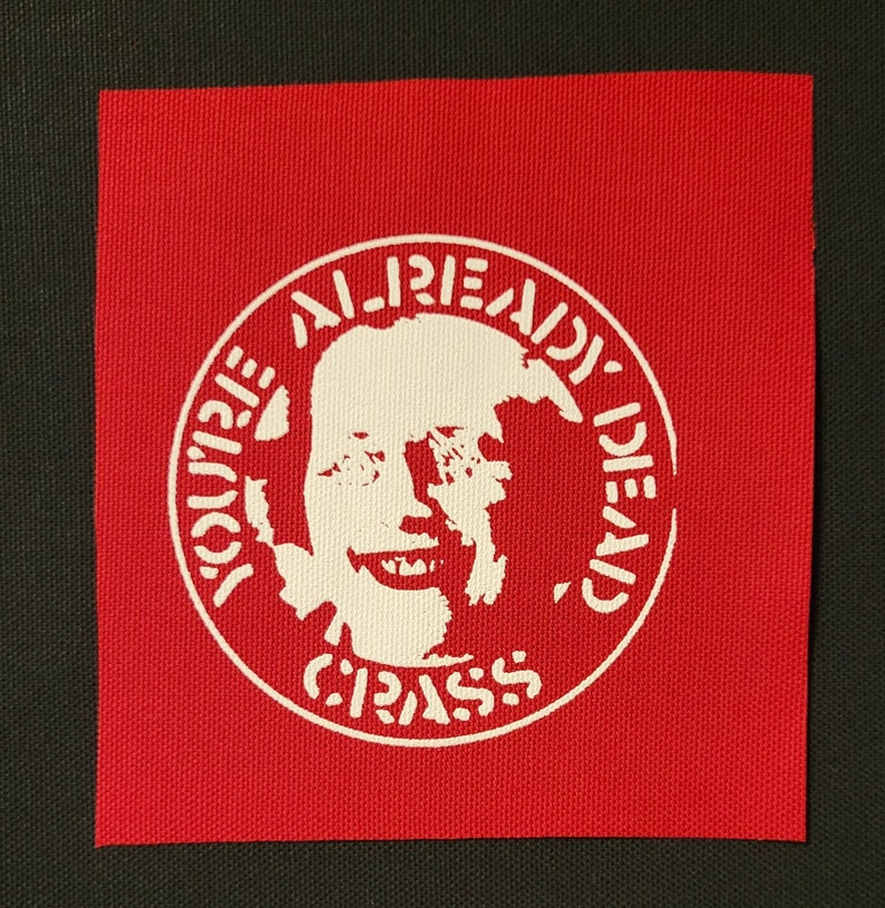 May include: A red fabric patch with a white circle containing a stylized image of a person's face and the text "YOU'RE ALREADY DEAD, CRASS".