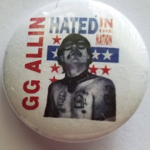 Puede incluir: Un botón redondo blanco con el texto "GG ALLIN" en rojo en el lado izquierdo. El botón presenta una imagen en blanco y negro de una persona con bigote y las palabras "HATED IN THE NATION" con estrellas y rayas.
