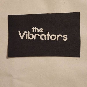 Peut inclure: Une étiquette rectangulaire noire avec le texte blanc "the Vibrators". L'étiquette est en tissu texturé et se détache sur un fond beige clair.