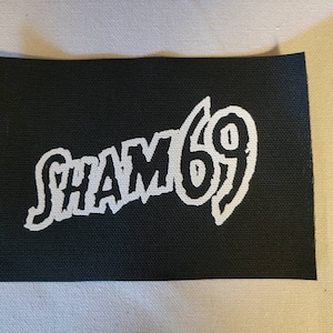 Puede incluir: Parche de tela negro con letras blancas que dicen "SHAM 69".