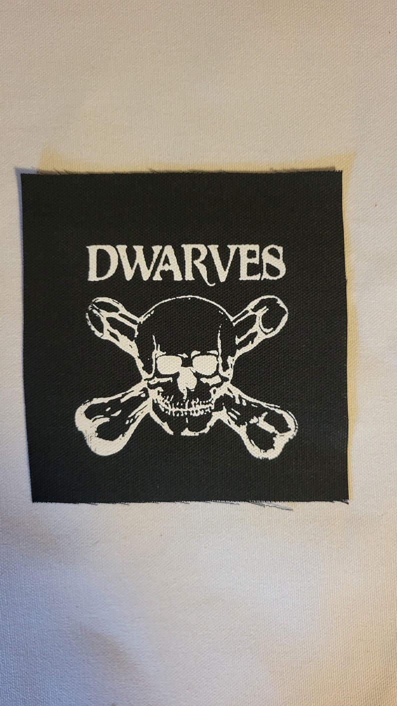 May include: Black and white embroidered patch featuring a skull and crossbones with the word "DWARVES" above it.