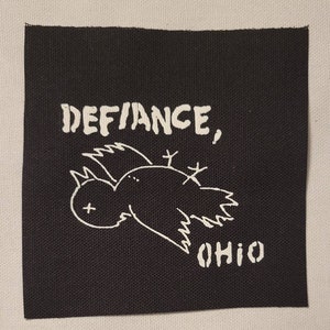 May include: Black square patch with white text and a bird illustration. The text reads "DEFANCE, OHIO". The bird is drawn in a simple, cartoonish style, with its wings spread and a cross mark on its eye.