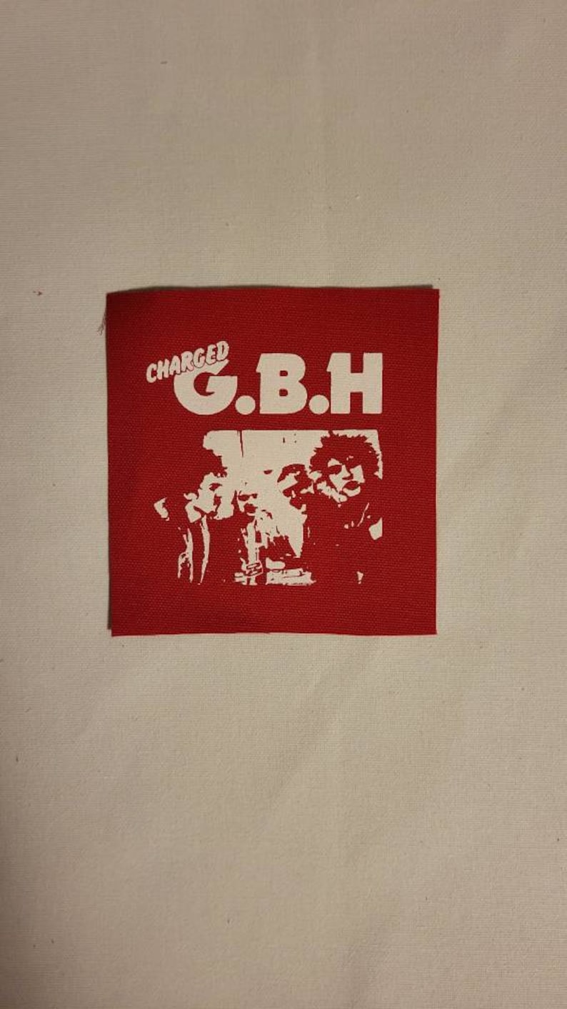 K&ouml;nnte beinhalten: Roter Stoff-Patch mit wei&szlig;em Text "CHARGED G.B.H." und einem Schwarzwei&szlig;bild einer Band.